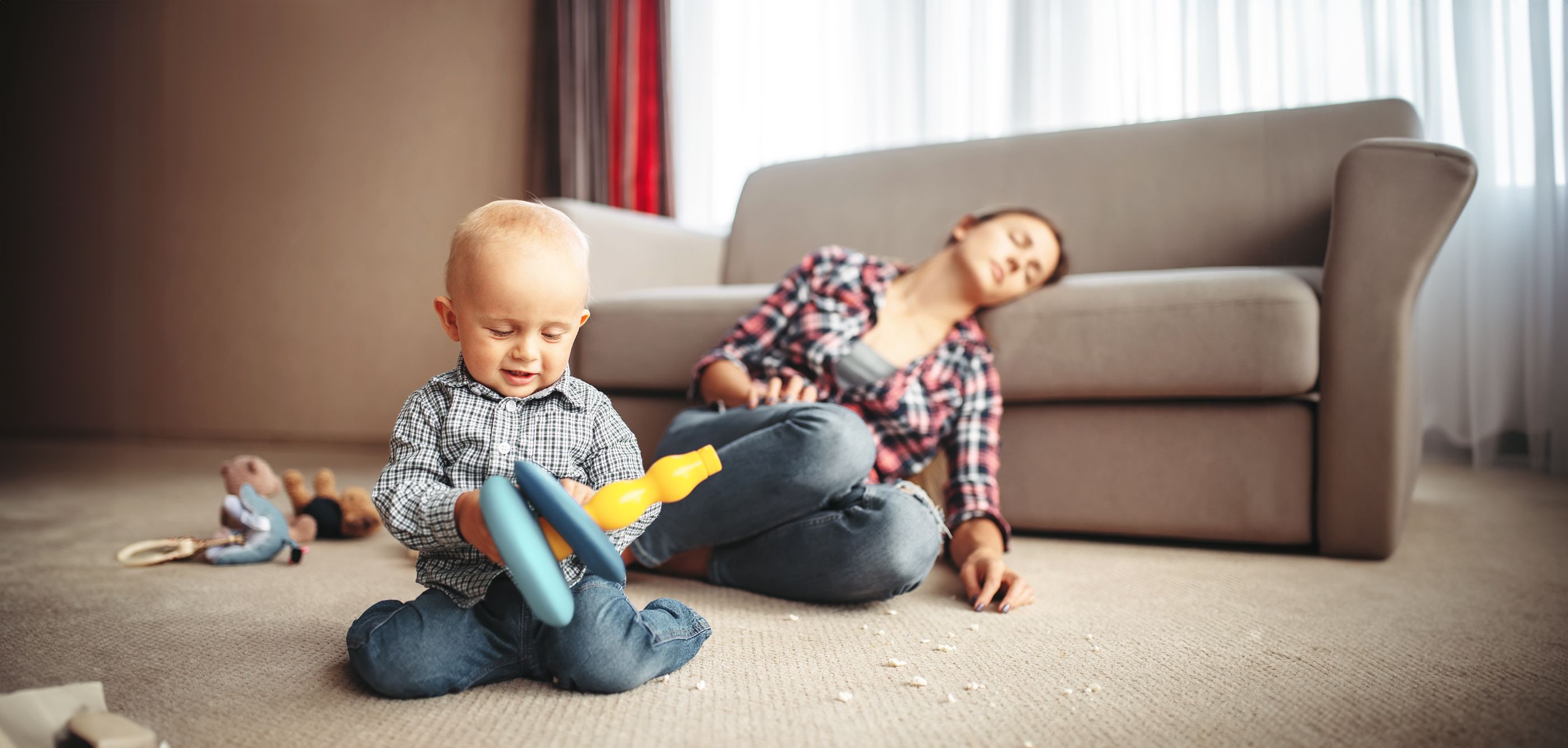 Baby spielt im Vordergrund und die Mutter sitzt erschöpft auf dem Boden und lehnt am Sofa